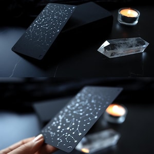 May include: Black tarot card with a constellation design in white. A clear quartz crystal and a lit candle are also visible. The card is held in a hand in one of the images. The background is dark.