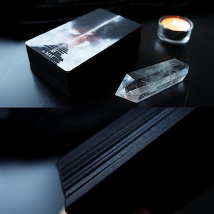 May include: A stack of black tarot cards with the text "LE MAT" on the top card. A clear quartz crystal and a lit candle are next to the cards. The top card features a stairway leading into a cloudy sky.