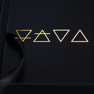 May include: A black box with gold geometric symbols. The symbols include a line with a downward-facing triangle, a triangle with a horizontal line, an inverted triangle, and a triangle. The symbols are arranged in a row.