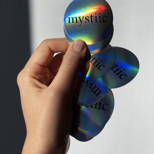 May include: A hand holding a stack of holographic round stickers. Each sticker has the word "mystiic" printed in black. The stickers have a rainbow sheen, reflecting various colors.