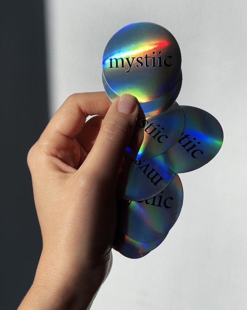 May include: A hand holding a stack of holographic round stickers. Each sticker has the word "mystiic" printed in black. The stickers have a rainbow sheen, reflecting various colors.