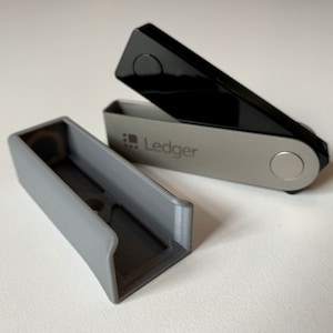 May include: A gray plastic case for a Ledger Nano S cryptocurrency hardware wallet. The case is open and the Ledger Nano S is visible inside. The Ledger Nano S is a small, rectangular device with a black screen and a silver button.