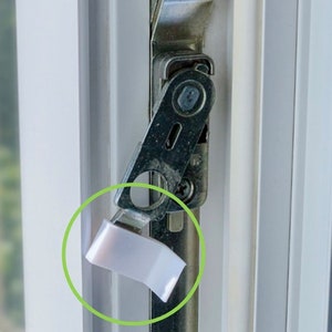 Peut inclure: Gros plan sur un loquet de fenêtre en plastique blanc sur un cadre de fenêtre blanc. Le loquet est fixé à un mécanisme métallique.
