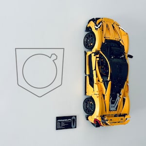 Könnte beinhalten: Ein gelbes und schwarzes Lego-Modell eines McLaren P1 ist an einer Wand montiert. Das Auto ist teilweise zerlegt, wodurch die komplizierten Details des Lego-Baus sichtbar werden. Das Auto wird mit einer schwarz-weißen Informationskarte mit der Aufschrift "McLaren P1" präsentiert.