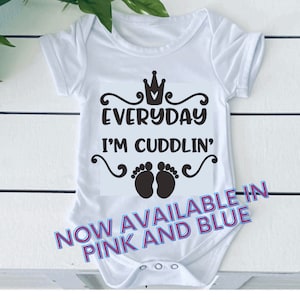 May include: White baby onesie with black text that reads "Everyday I'm Cuddlin'" with a crown and two footprints. The text "NOW AVAILABLE IN PINK AND BLUE" is written in pink and blue on the bottom of the onesie.
