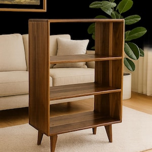 May include: A mid-century modern bookshelf made of dark wood. The bookshelf has three shelves and a rectangular frame. The legs are angled. A painting of a landscape hangs on the wall behind the bookshelf.