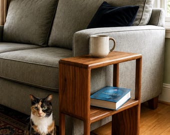 Puede incluir: Una mesa auxiliar rectangular de madera con dos estantes abiertos, con un acabado de veta de madera natural lisa. Una taza de cerámica beige descansa sobre la superficie superior y un libro de cubierta azul se sitúa en el estante intermedio.