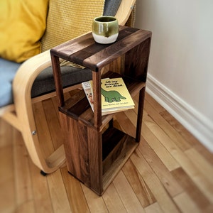 May include: A wooden side table with two shelves, a book titled "All my friends are dead" on the top shelf, and a green ceramic mug on the top shelf. The table is made of walnut wood and has a natural finish.
