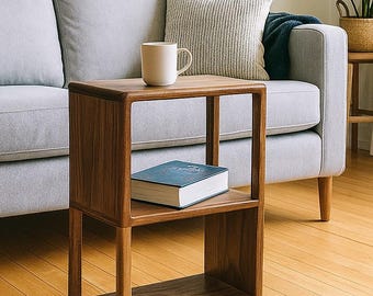 Könnte beinhalten: Ein moderner Beistelltisch aus Holz mit zwei Regalen, auf dem sich eine weiße Tasse und ein blaues Buch befinden. Der Tisch steht neben einem grauen Sofa mit einem dekorativen Kissen. Ein gerahmtes Landschaftskunstwerk hängt an der Wand über dem Sofa.