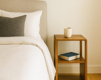 Puede incluir: Interior de dormitorio con cama, edredón blanco y cojín gris. Una mesita de noche de madera con una taza y un libro. Una obra de arte abstracta cuelga de la pared. Una lámpara colgante cuelga del techo.