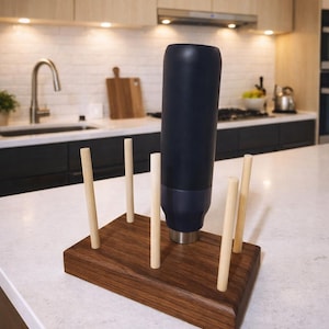 May include: A navy blue water bottle in a wooden holder with six wooden dowels. The holder is made of dark brown wood and is rectangular. The bottle is on a white countertop in a kitchen setting.