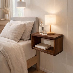May include: A dark wood floating bedside table with a lamp, a glass of water, and a bottle. A book rests on the shelf. The bed has a grey headboard and light-coloured bedding. A pendant light hangs above.