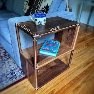 Puede incluir: Una mesa auxiliar de madera de nogal con dos estantes. La mesa mide 46 cm de ancho, 41 cm de profundidad y 61 cm de alto. La mesa tiene una taza azul con un dise&ntilde;o de hongo en el estante superior y un libro titulado "Alice's Adventures in Wonderland" en el estante inferior.