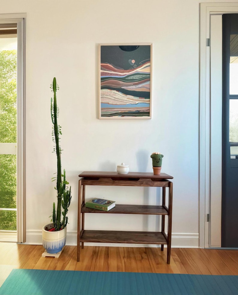May include: A dark wood console table with three shelves. The table has a white ceramic jar on the top shelf, a small green cactus in a terracotta pot on the top shelf, and a book on the middle shelf. The table is in front of a white wall with a framed abstract print.