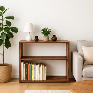 K&ouml;nnte beinhalten: Ein rechteckiges B&uuml;cherregal aus Holz mit zwei offenen F&auml;chern. Auf der oberen Ablage stehen eine wei&szlig;e Tischlampe, eine kleine gr&uuml;ne Pflanze in einem beigen Topf und zwei dunkelbraune Vasen. Das untere Fach enth&auml;lt eine Reihe von B&uuml;chern.