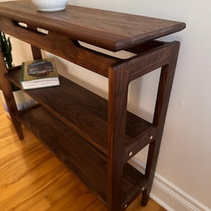 May include: A dark brown wooden console table with two shelves and a top shelf. The table has a unique design with a curved top and angled legs. The table is made of solid wood and has a natural finish.