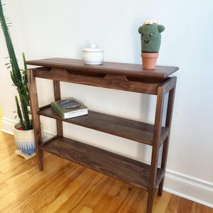 May include: A dark brown wooden console table with two shelves and a top shelf. The table has a simple design with straight lines and rounded edges. A small white ceramic jar and a green crocheted cactus are on the top shelf. A book is on the lower shelf.