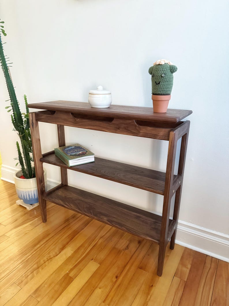 May include: A dark brown wooden console table with two shelves and a top shelf. The table has a simple design with straight lines and rounded edges. A small white ceramic jar and a green crocheted cactus are on the top shelf. A book is on the lower shelf.