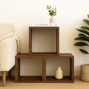 Peut inclure: Deux étagères cubiques en bois brun foncé empilées, avec un vase crème et un livre intitulé "THE KINFOLK HOME" sur le dessus. Un petit vase avec de la verdure et un panier en osier sont également visibles.