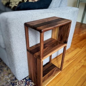Puede incluir: Una mesa auxiliar de madera de dos niveles con acabado en nogal. La mesa tiene un dise&ntilde;o simple y rectangular con estantes abiertos. La mesa est&aacute; colocada junto a un sof&aacute; gris.