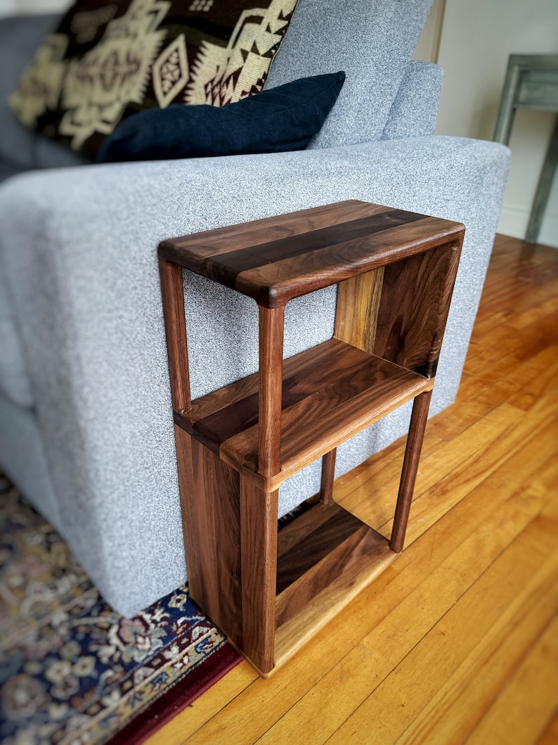 Puede incluir: Una mesa auxiliar de madera de dos niveles con acabado en nogal. La mesa tiene un dise&ntilde;o simple y rectangular con estantes abiertos. La mesa est&aacute; colocada junto a un sof&aacute; gris.