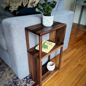 Puede incluir: Una mesa auxiliar de madera de nogal de dos niveles con una peque&ntilde;a planta de cactus en maceta, un libro titulado "All My Friends are Dead" y un portavelas blanco en las estanter&iacute;as. La mesa est&aacute; unida a un sof&aacute; gris.
