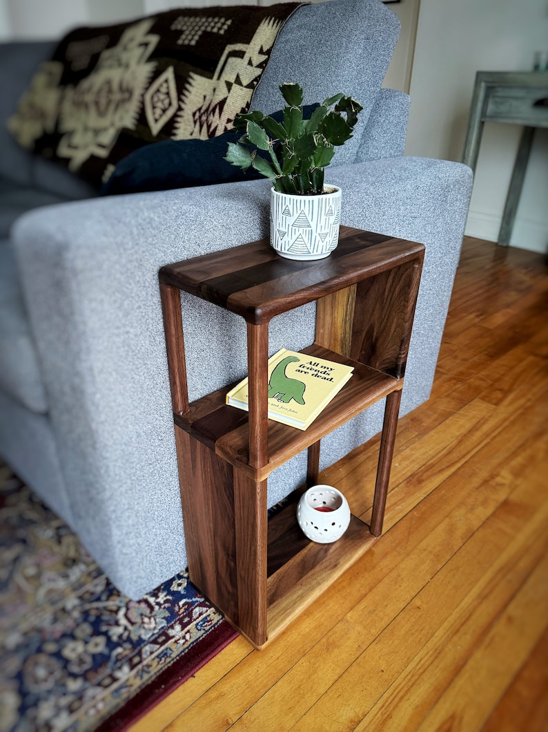 Puede incluir: Una mesa auxiliar de madera de nogal de dos niveles con una peque&ntilde;a planta de cactus en maceta, un libro titulado "All My Friends are Dead" y un portavelas blanco en las estanter&iacute;as. La mesa est&aacute; unida a un sof&aacute; gris.