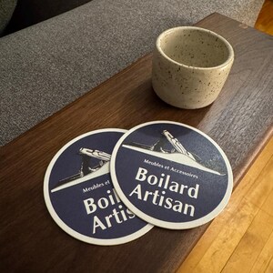 Pu&ograve; includere: Due sottobicchieri rotondi blu e bianchi con la scritta "Meubles et Accessoires Boillard Artisan" stampata su di essi. I sottobicchieri sono su un tavolo di legno accanto a una tazza di ceramica bianca macchiata.