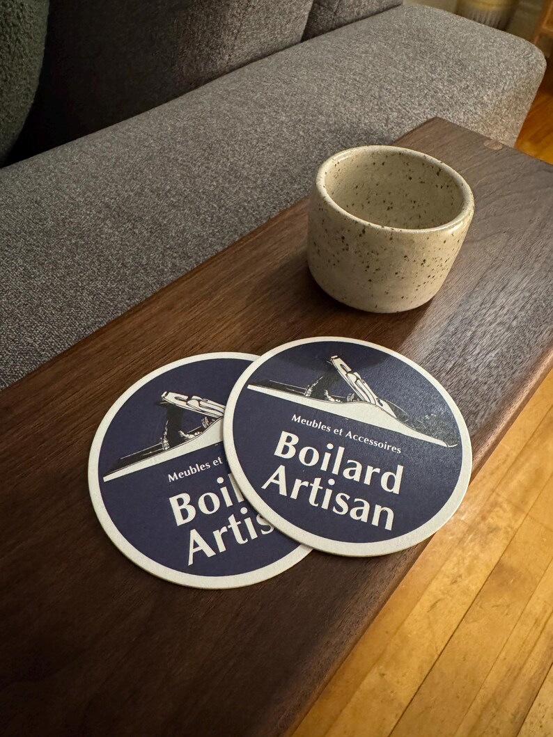 Pu&ograve; includere: Due sottobicchieri rotondi blu e bianchi con la scritta "Meubles et Accessoires Boillard Artisan" stampata su di essi. I sottobicchieri sono su un tavolo di legno accanto a una tazza di ceramica bianca macchiata.