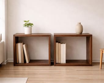 Può includere: Due scaffali aperti quadrati in legno marrone scuro posizionati fianco a fianco. L'unità di sinistra contiene una piccola pianta verde in un vaso bianco e tre libri. L'unità di destra contiene un vaso in ceramica beige liscio e tre libri.