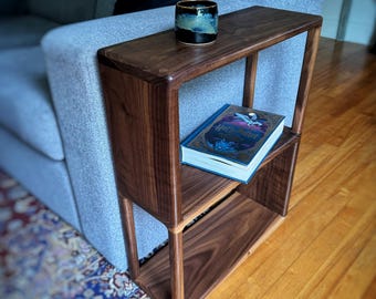 Puede incluir: Una mesa auxiliar de madera marrón oscuro con dos estantes. Una taza de cerámica con detalles azules y dorados está en el estante superior. Un libro con una cubierta azul está en el estante inferior. La mesa está junto a un sofá gris.