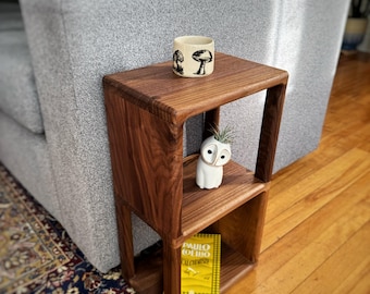 Könnte beinhalten: Ein zweistufiger Beistelltisch aus Holz mit quadratischer Oberseite und offenen Regalen. Der Tisch ist aus dunklem Walnussholz gefertigt und hat eine natürliche Oberfläche. Eine kleine weiße Keramiktasse mit einem schwarzen Pilzdesign steht auf dem oberen Regal. Eine kleine weiße Eulenfigur steht auf dem unteren Regal. Ein gelbes Taschenbuch mit dem Titel "Paulo Coelho, Der Alchemist" liegt auf dem unteren Regal.