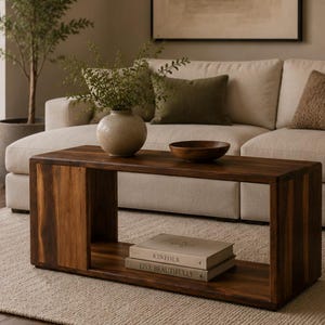 May include: A rectangular dark wood coffee table with an open shelf design. The top surface holds a speckled beige ceramic vase with green foliage and a small dark wooden bowl. Two stacked books titled Kinfolk and Live Beautifully sit below.