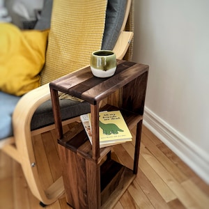 Puede incluir: Una mesa auxiliar de madera con dos estantes, un libro titulado "All my friends are dead." y una taza de cer&aacute;mica verde en el estante superior. La mesa est&aacute; junto a una manta amarilla en una silla.