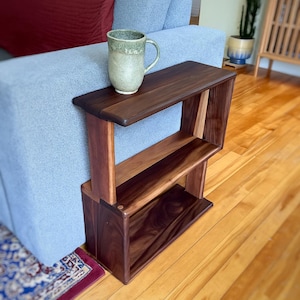 May include: A handcrafted wooden side table with two shelves. The table is made of dark walnut and lighter wood, with a rectangular top and a sturdy, open design. A ceramic mug with a green glaze sits on the top shelf. The table is 24 inches tall.