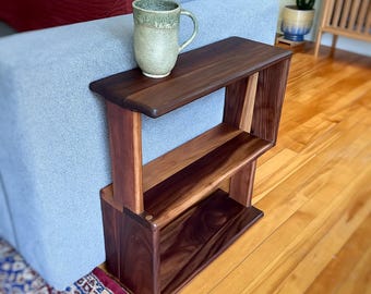 May include: A dark brown wooden side table with a rectangular top, a middle shelf, and a solid base, featuring a smooth finish and visible wood grain. A speckled, light green ceramic mug with a handle sits on the top surface of the table.