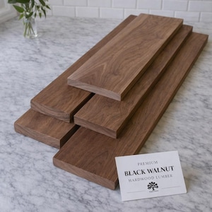 Op de afbeelding: Verschillende rechthoekige planken van donkerbruin zwart walnoothout liggen op een marmeren oppervlak. Een witte kaart met de tekst "Premium Black Walnut Hardwood Lumber" is zichtbaar. Een vaas met witte bloemen staat op de achtergrond. De planken zijn ongeveer 76 cm lang.