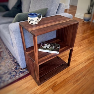 May include: A dark walnut side table with two shelves and a top surface. A ceramic mug with a mushroom design sits on top. A book is on the middle shelf. The table is next to a gray sofa.