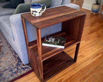 May include: A dark walnut side table with two shelves and a top surface. A ceramic mug with a mushroom design sits on top. A book is on the middle shelf. The table is next to a gray sofa.