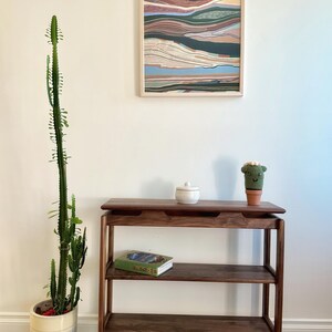 May include: A dark wood console table with three shelves. The top shelf has a small white ceramic jar and a small green crocheted cactus. The middle shelf has a green book with a blue cover. The bottom shelf is empty. The table is in front of a white wall with a framed abstract painting.