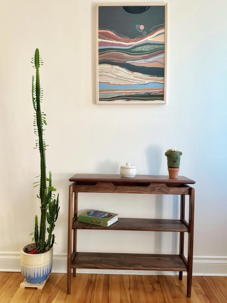 May include: A dark wood console table with three shelves. The top shelf has a small white ceramic jar and a small green crocheted cactus. The middle shelf has a green book with a blue cover. The bottom shelf is empty. The table is in front of a white wall with a framed abstract painting.