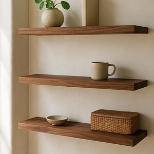 May include: Three dark wood floating shelves mounted on a light-colored wall. The top shelf holds a small plant in a beige pot and a book. The middle shelf has a beige mug. The bottom shelf has a small bowl and a woven basket.
