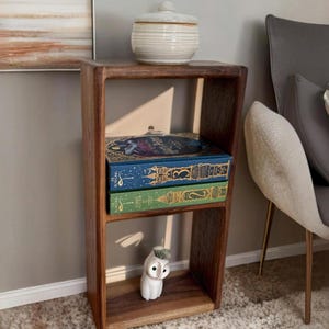May include: A wooden shelf unit with two shelves, featuring a white ceramic owl figurine, a white ceramic jar with a lid, and three books with colourful covers. The shelf is against a light beige wall.