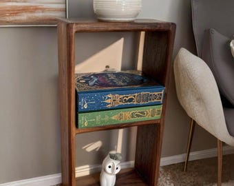 May include: A wooden shelf unit with two shelves, featuring a white ceramic owl figurine, a white ceramic jar with a lid, and three books with colorful covers. The shelf is against a light beige wall.