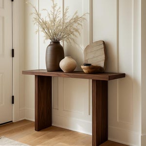 May include: A dark wood console table with a minimalist design. It features a rectangular top and two thick, rectangular legs. On the table are a large brown vase with dried plants, a small beige vase, a wooden bowl, and a decorative wooden piece.