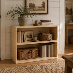 Puede incluir: Una estantería de madera clara con dos estantes abiertos. El estante superior contiene un jarrón de cerámica con vegetación, una vela pequeña, un cuenco de madera y libros. El estante inferior contiene una cesta tejida y varios libros.