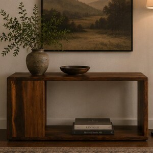 May include: A rectangular dark wood console table with a solid side panel and an open shelf. On the top surface sits a textured gray vase with green foliage and a dark brown bowl. Two stacked books are placed on the lower shelf of the table.