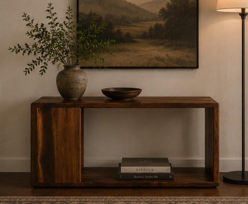 May include: A rectangular dark wood console table with a solid side panel and an open shelf. On the top surface sits a textured gray vase with green foliage and a dark brown bowl. Two stacked books are placed on the lower shelf of the table.