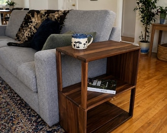 May include: A dark brown wooden side table with two shelves. The table is rectangular and has a modern design. A mug with a mushroom design sits on top. The table is next to a gray sofa.