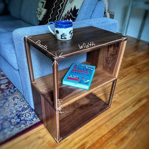 Pu&ograve; includere: Un tavolino da salotto in legno scuro con due ripiani. Il tavolino &egrave; largo 46 cm, profondo 41 cm e alto 61 cm. Una tazza blu con un motivo a fungo si trova sul ripiano superiore. Un libro intitolato "Alice's Adventures in Wonderland" si trova sul ripiano inferiore.